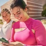 Mulher vestindo roupa de treino rosa sorri enquanto usa o smartphone ao ar livre, acompanhada por colegas também com roupas esportivas.