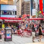 Vista de uma praça movimentada em uma cidade dos Estados Unidos, com pedestres caminhando, mesas vermelhas ao ar livre e prédios altos com outdoors comerciais ao fundo
