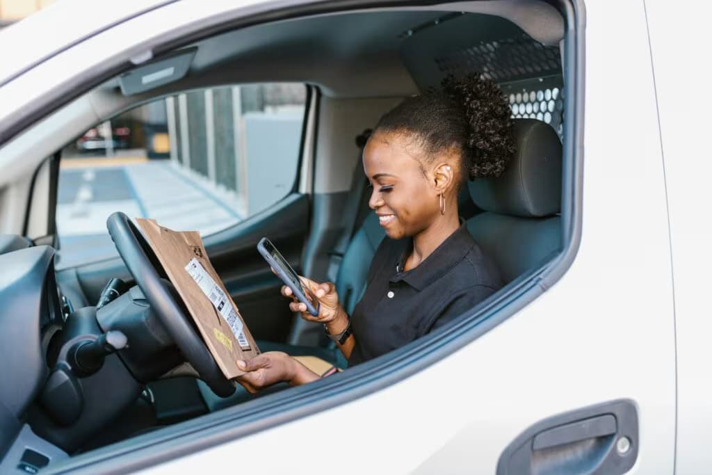 Mulher negra sorridente dentro de uma van de entregas, segurando um pacote de papel pardo e utilizando um smartphone para conferir os dados da entrega da Nuvemshop.