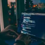 Pessoa programando em um laptop durante a noite, com luzes da cidade ao fundo refletidas na janela, ilustrando o conceito de vibe coding.